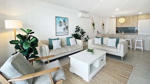 Un salon moderne avec deux canapés, une table basse et un fauteuil. On aperçoit, au fond, un coin cuisine avec un plan de travail et des plantes suspendues.