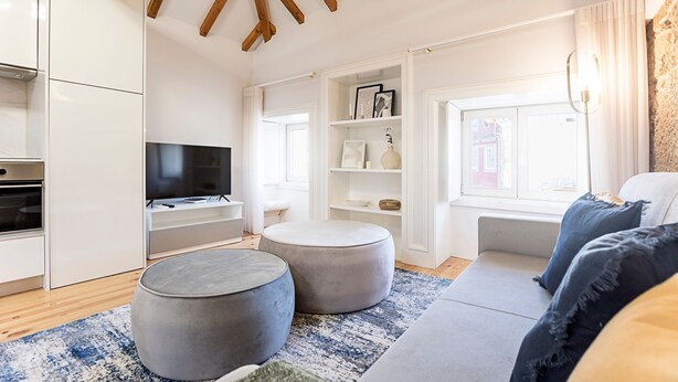 Un salon moderne doté d’un téléviseur à écran plat, de deux poufs, d’un canapé avec des coussins bleus et d’une fenêtre offrant une vue agréable.