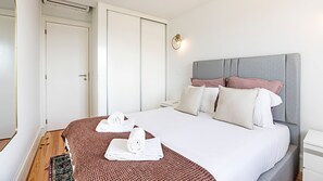 A hotel room with a bed, a bedside table, a nightstand, a wall-mounted light, and a door.