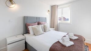 Une chambre à coucher moderne avec une tête de lit grise, du linge de lit blanc et une vue sur les bâtiments par la fenêtre.