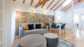 Un salon moderne avec un mur en pierre, un canapé gris agrémenté de coussins, une table à manger avec des chaises bleues, et un coin cuisine à l’arrière-plan.