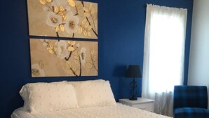 A bedroom with a large bed, a nightstand, a lamp, a chair, and two wall-mounted paintings.