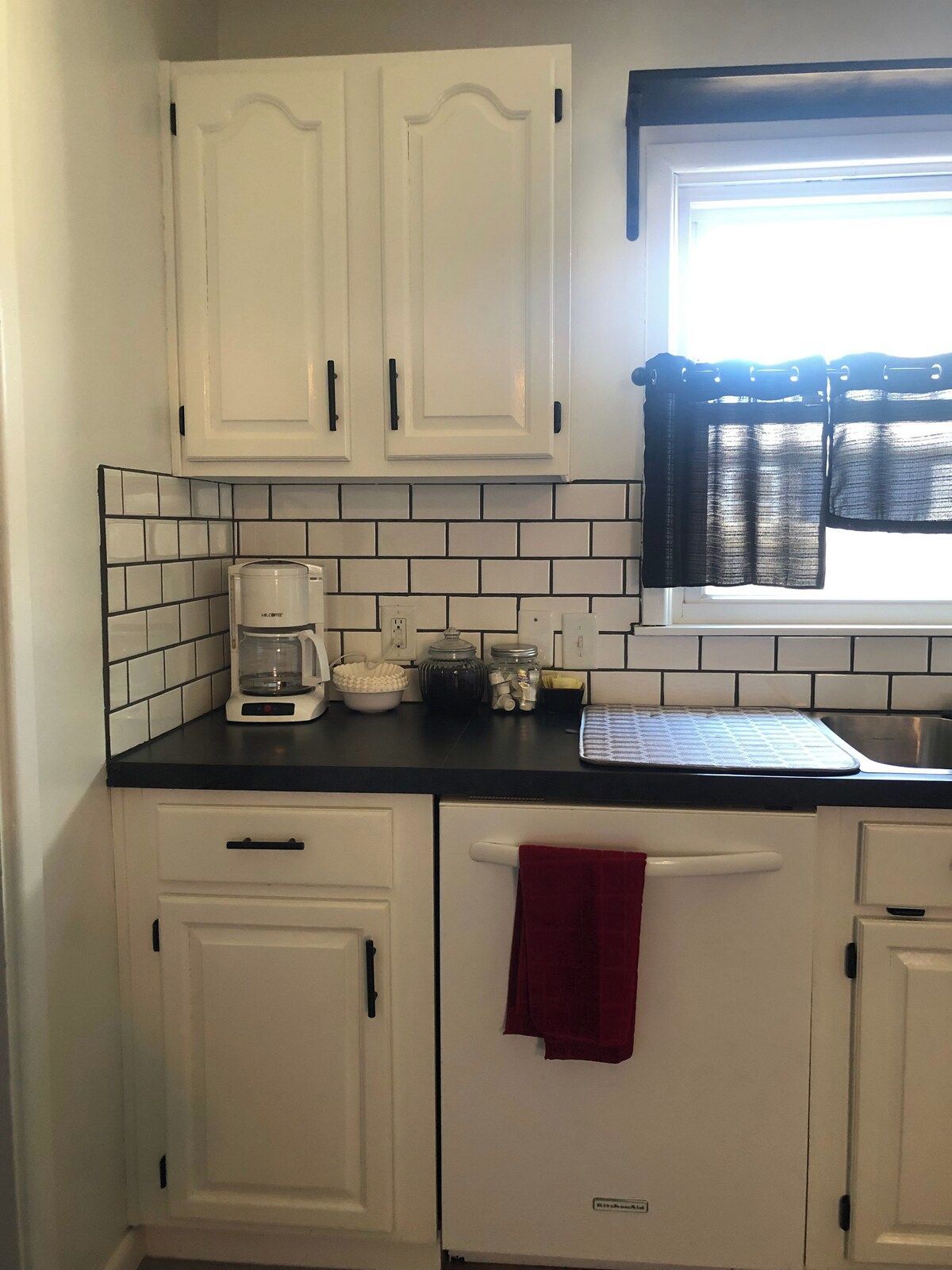 A kitchen with white cabinets, a coffee maker, a kettle, and a window with a curtain.