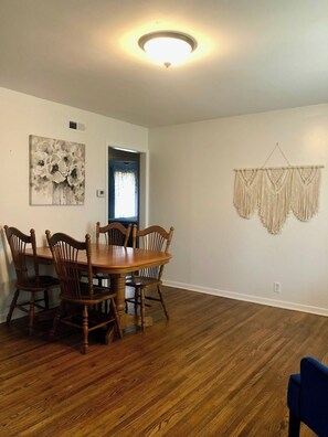 Un coin repas avec une table et des chaises en bois, un luminaire au plafond, une décoration murale en macramé et une fenêtre avec des rideaux.