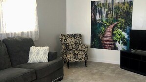 Un soggiorno con un divano grigio scuro, una poltrona fantasia, una ventola a soffitto, un quadro di un sentiero boschivo e un televisore su un mobile.