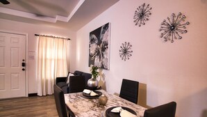Un área de comedor con una mesa de mármol, sillas negras y un florero con flores. Se observan decoraciones en la pared y una puerta al fondo.