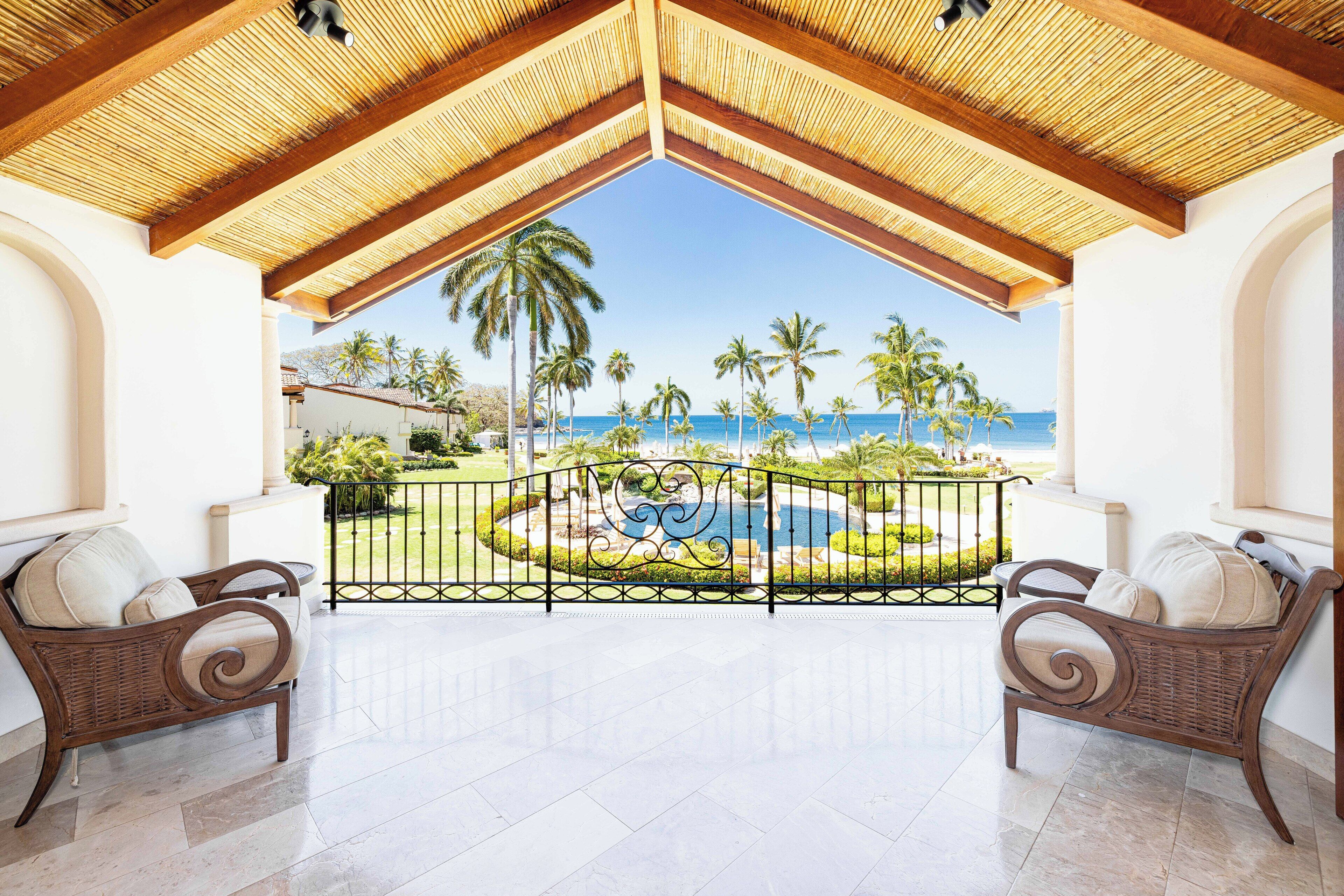 A balcony with a view of the beach, two wicker chairs, and a decorative fence.
