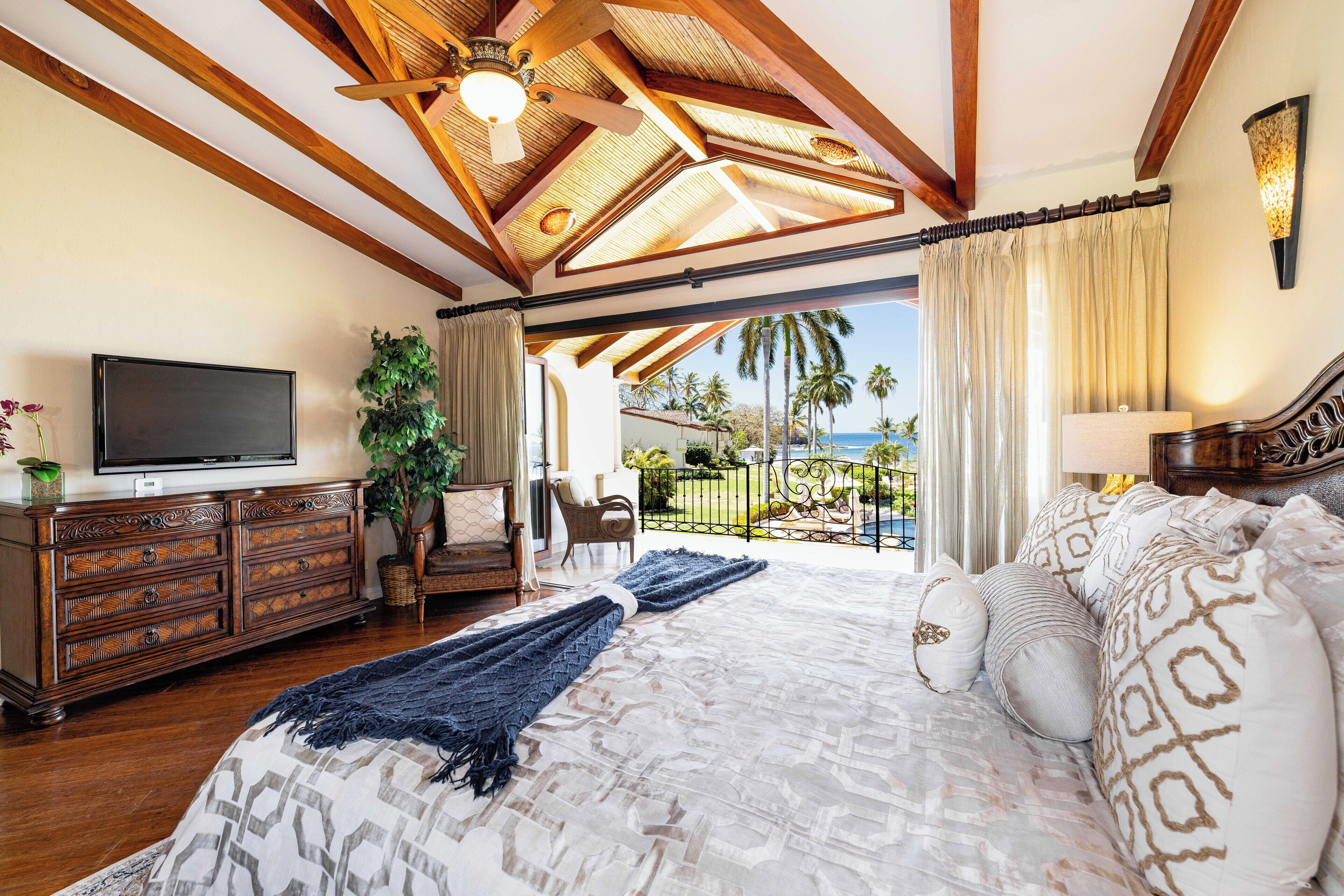 A spacious bedroom with a large bed, wooden ceiling beams, a television, and a view of the outdoors.