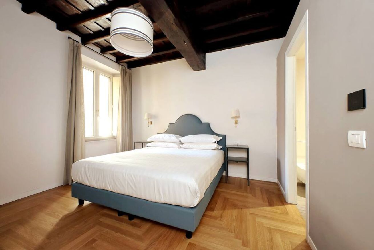 A bedroom with a bed, wooden flooring, a window with curtains, a ceiling light fixture, and a bedside table with a lamp.