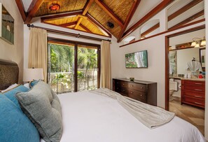 A bedroom with a large bed, a wooden ceiling, a balcony with a view of greenery, a dresser, and a bathroom with a mirror and sink. - Stay 24 - Newly Renovated Beachfront Villa at The Palms Playa Flamingo. (Playa Flamingo)