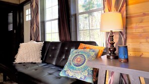 A room with a black leather sofa, a patterned cushion, a table with a lamp, and a window with curtains.