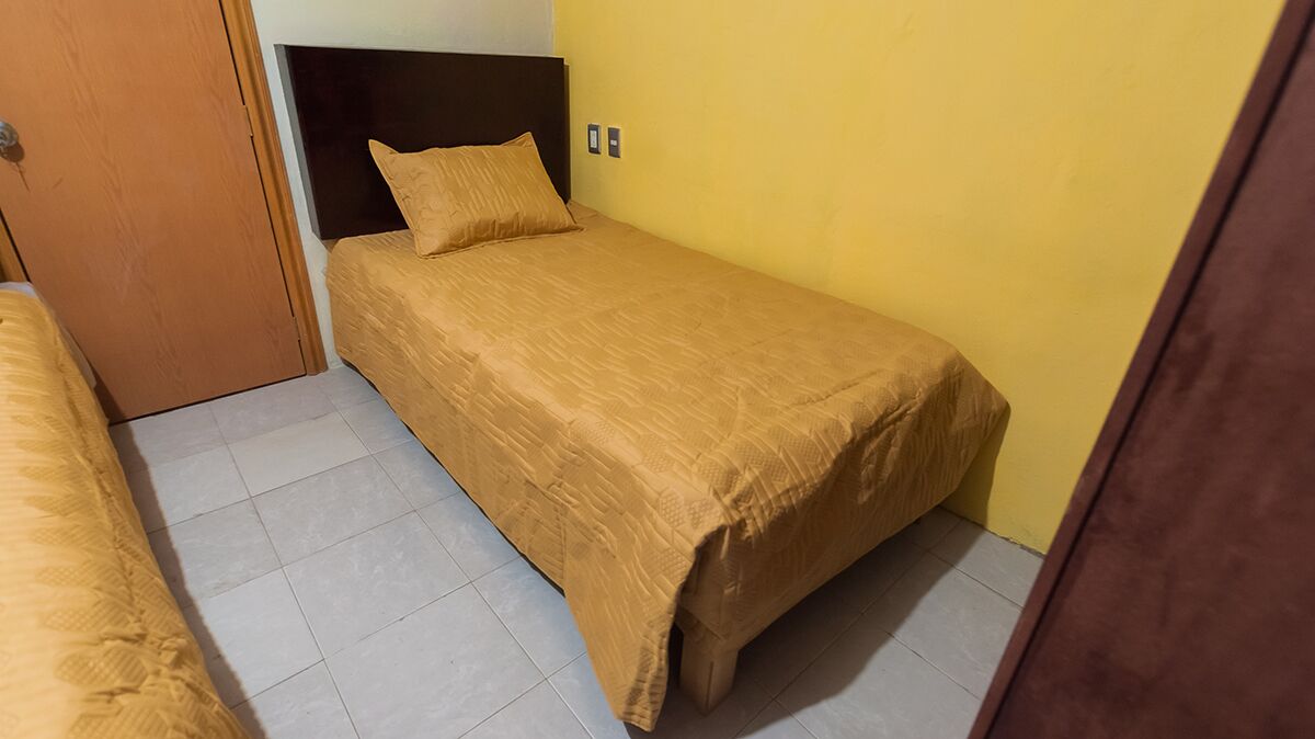 A small, single-bed room with a wooden headboard, a yellow wall, and a tiled floor.