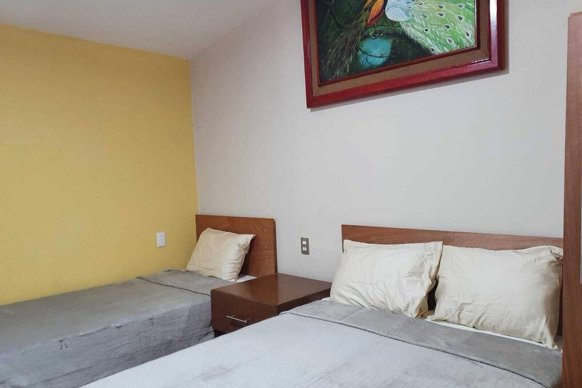 Ein Zimmer mit zwei Betten, jeweils mit einem hölzernen Kopfteil und einer grauen Tagesdecke. Dazwischen steht ein kleiner Nachttisch aus Holz, und ein Gemälde hängt über den Betten an der Wand.