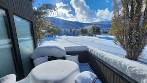 Un patio cubierto de nieve con una mesa y sillas, una cerca de madera y vistas a un paisaje nevado con árboles y montañas al fondo.