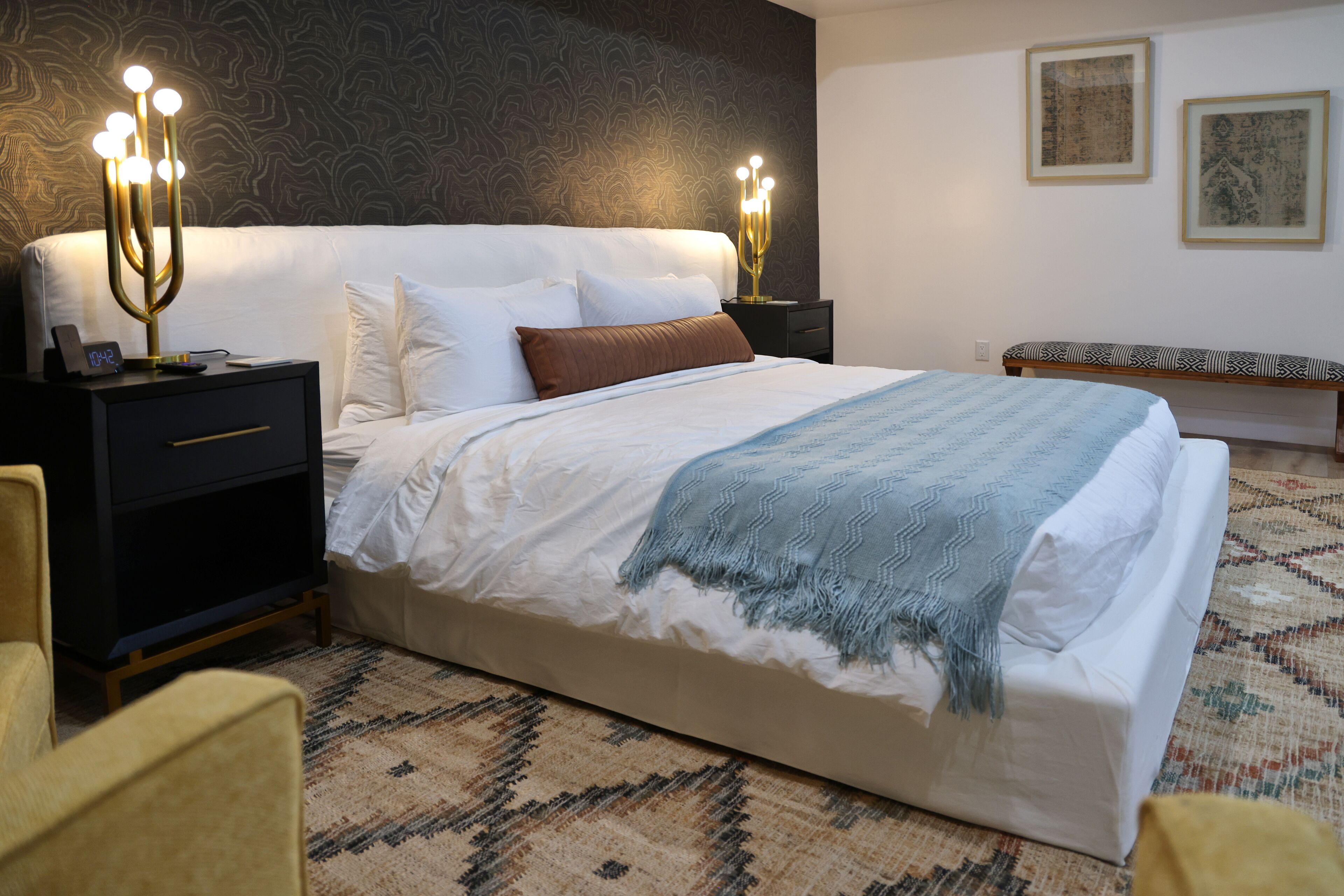 A neatly made bed with a white comforter and a blue throw blanket, flanked by two bedside tables with lamps, a nightstand with a clock, and a patterned rug on the floor.