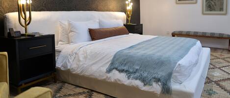 A neatly made bed with a white comforter and a blue throw blanket, flanked by two bedside tables with lamps, a nightstand with a clock, and a patterned rug on the floor.