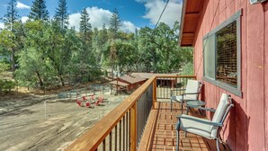 A wooden deck with chairs and a table, surrounded by a red building and a forested area. - Pet-Friendly Escape w/ Game Room & Fire Pit! (North Fork)