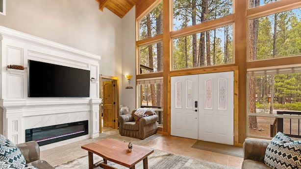 A spacious living room with a fireplace, a large TV, and a view of the forest through large windows.