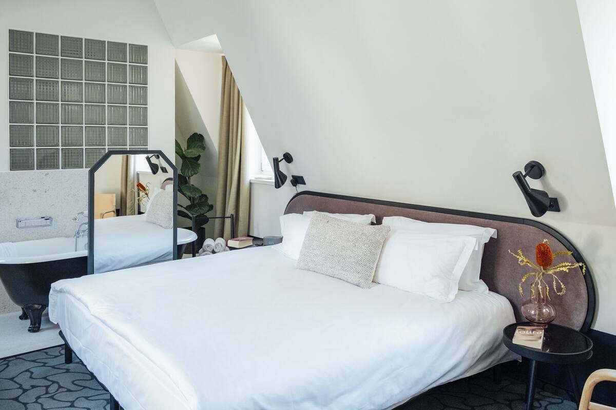 A modern hotel room with a large bed, a separate bathroom with a bathtub, a small table with a vase, and a large window with curtains.