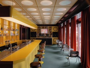 Un espace bar avec un comptoir jaune, de nombreux tabourets, un téléviseur et une bibliothèque contenant divers objets.