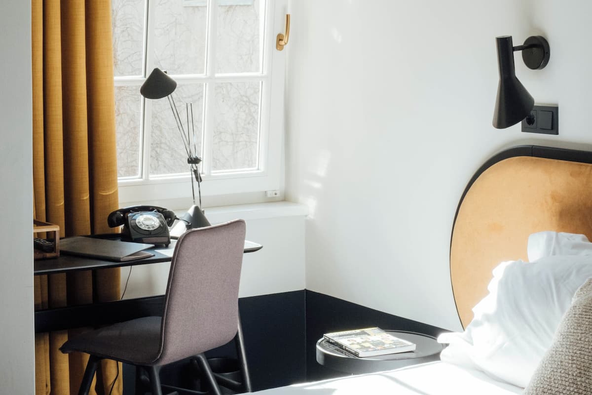 A hotel room with a bed, a desk with a chair, a lamp, and a window with curtains.