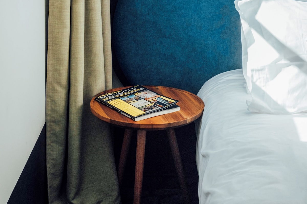 A hotel room with a blue headboard, a small wooden side table with a magazine, beige curtains, and a wall-mounted light fixture.