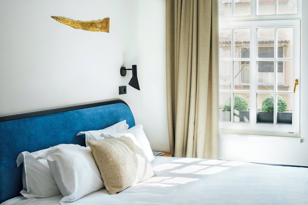 A hotel room with a blue headboard, white bedding, and a view of a building through a window with beige curtains.