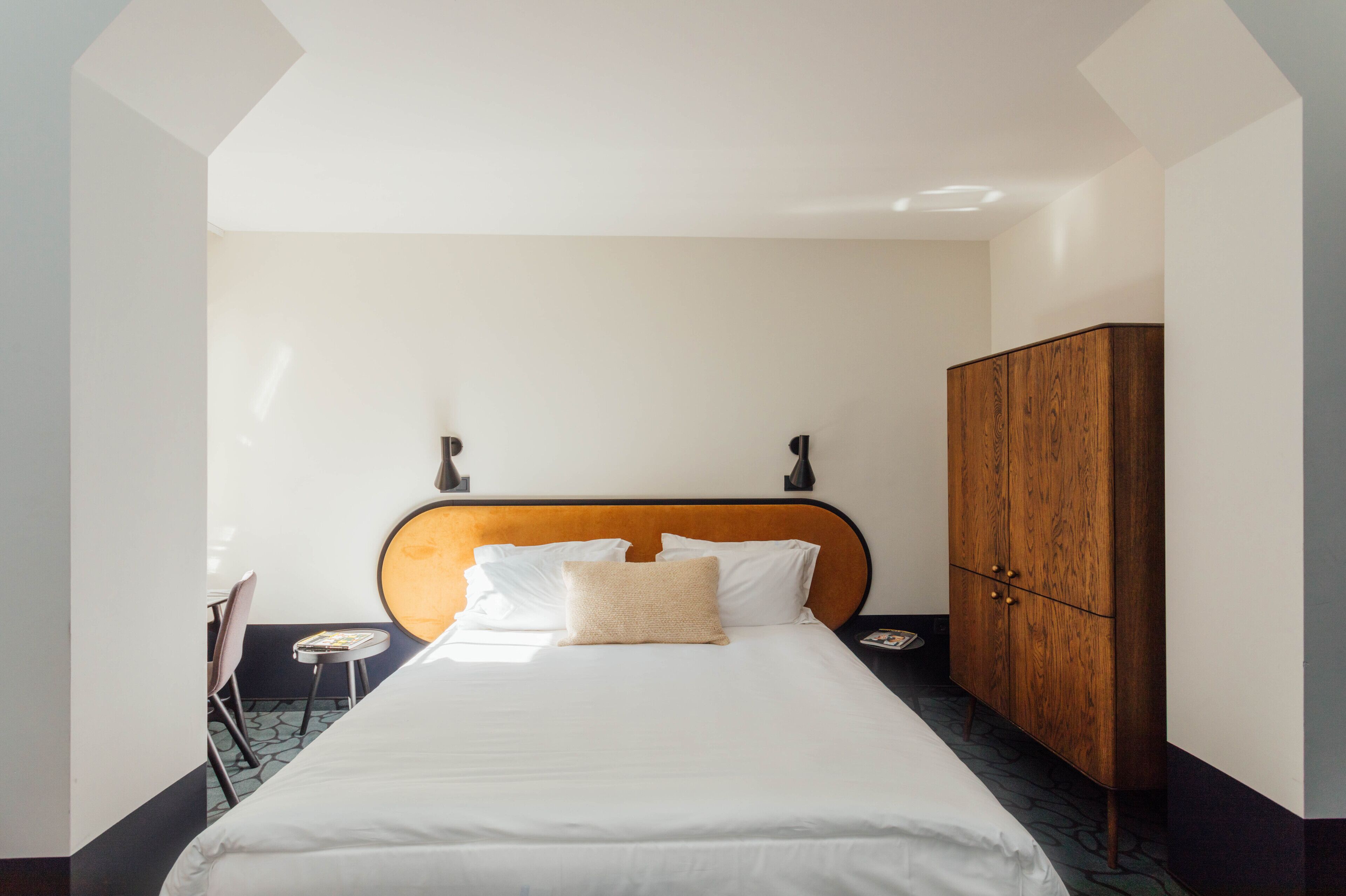 A hotel room with a bed, a wooden wardrobe, a small table with a chair, and a wall-mounted lamp.