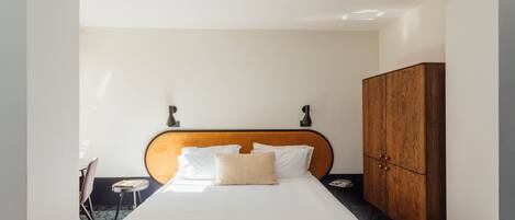 A hotel room with a bed, a wooden wardrobe, a small table with a chair, and a wall-mounted lamp.