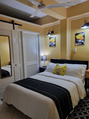 Una habitación de hotel con cama, ventilador de techo, mesita de noche con lámpara y dos cuadros en la pared.