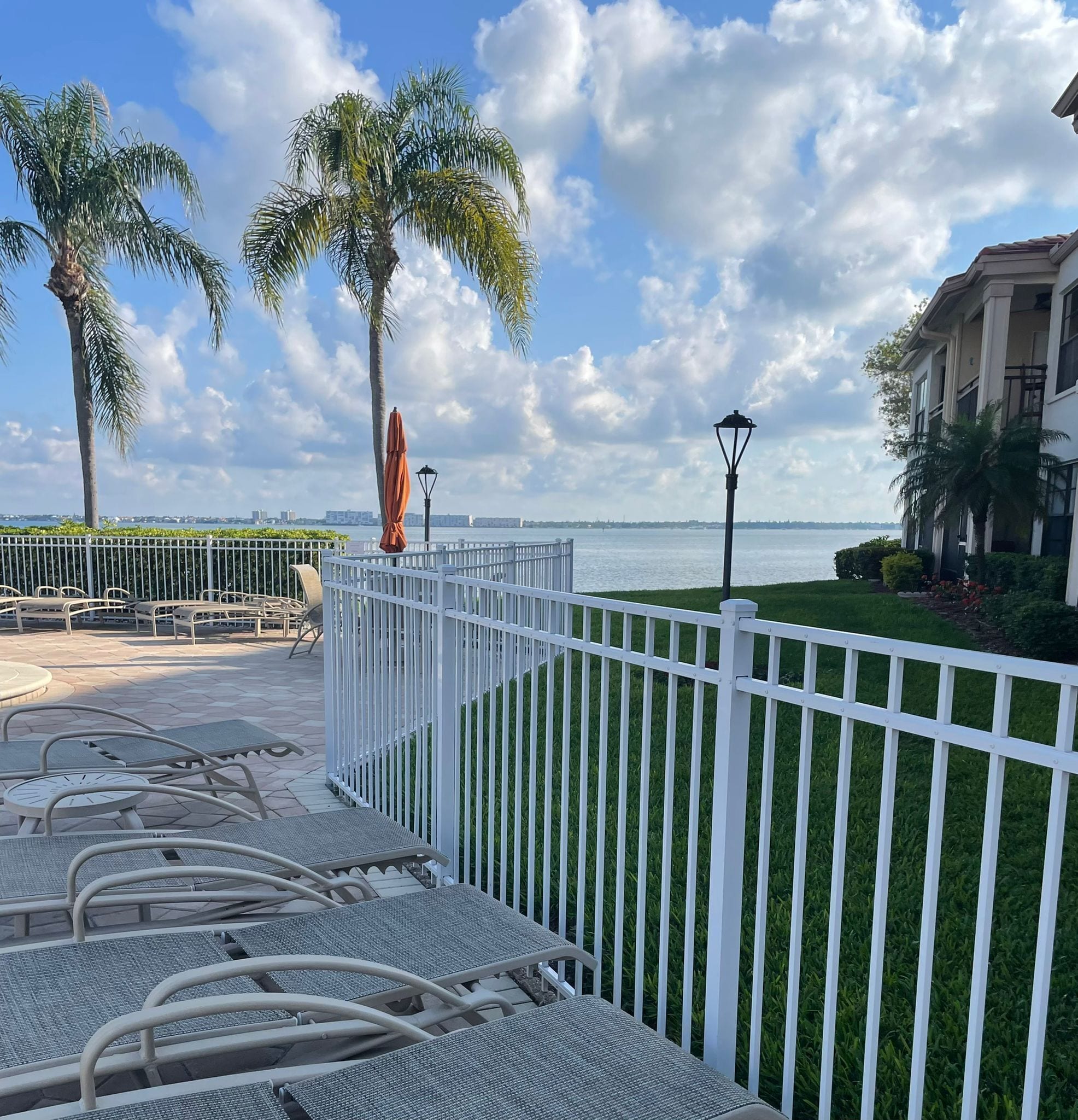 A waterfront patio with metal chairs and tables, a white fence, and palm trees.
