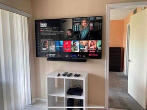 A flat-screen TV mounted on a wall with a Netflix interface displaying various shows. Below the TV, there is a white entertainment unit with electronic devices.