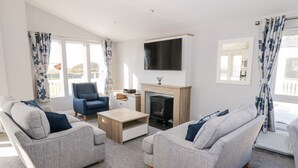 A modern living room with grey sofas, a blue armchair, a wooden coffee table, a fireplace, and a flat-screen TV. - Dershel Lodge (Dumfries)