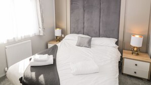 A neatly made bed with white linens and a grey headboard, two bedside tables with lamps, and a window with sheer curtains. - Dershel Lodge (Dumfries)