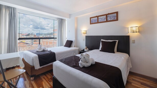 A hotel room with two beds, a table, a chair, and a view of the cityscape. - kAPAC INN HOTEL (Cusco)