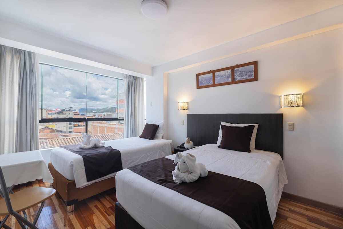 A hotel room with two beds, a table, a chair, and a view of the cityscape.