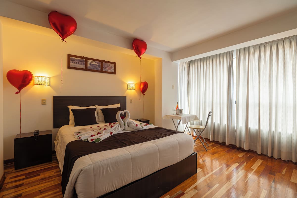 A hotel room with a large bed, a bedside table, a small table with a chair, and a window with curtains.
