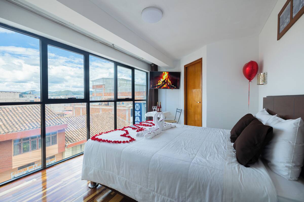 A hotel room with a large window, a bed with white bedding, a red balloon, and a television on the wall.