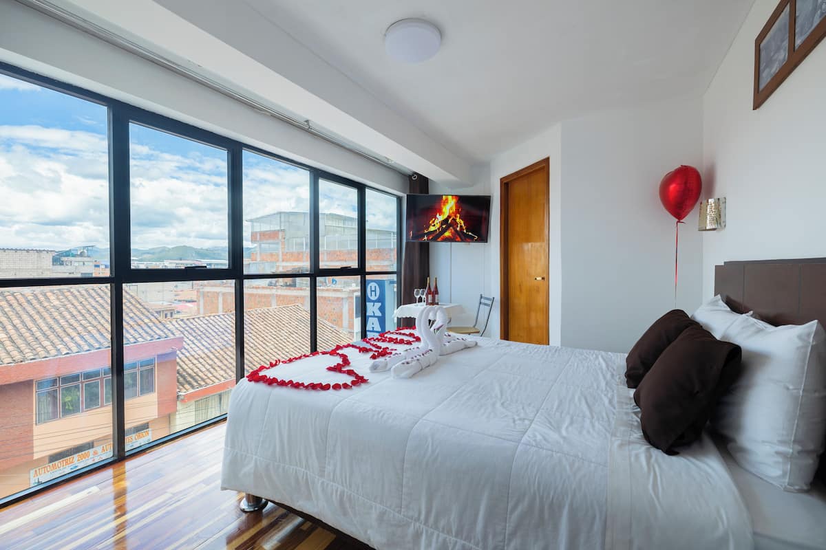 Quarto de hotel com uma janela grande, uma cama com roupa de cama branca, um balão vermelho e uma televisão na parede.