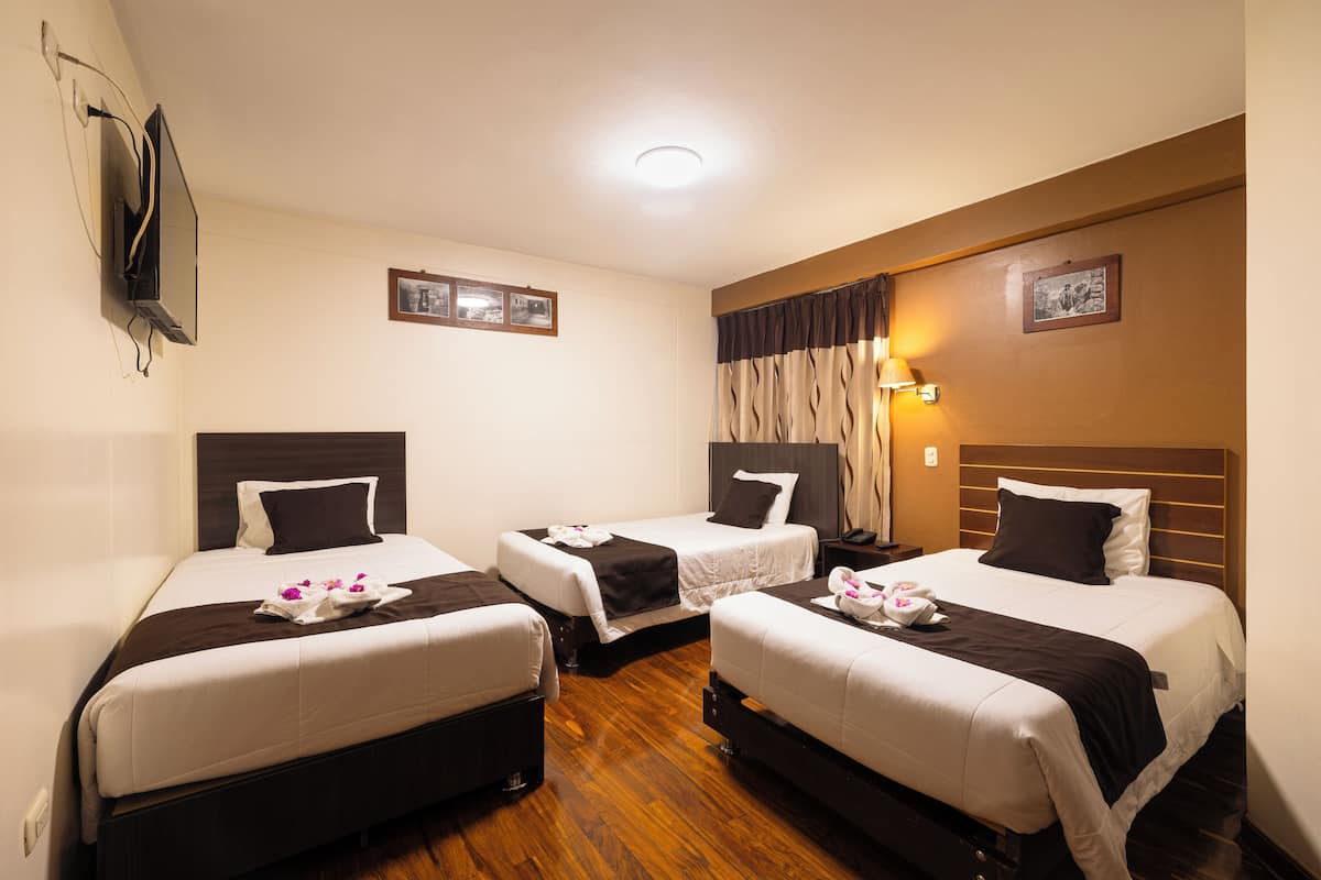 A hotel room with three single beds, a wooden floor, a flat-screen TV mounted on the wall, and a small table with a lamp.