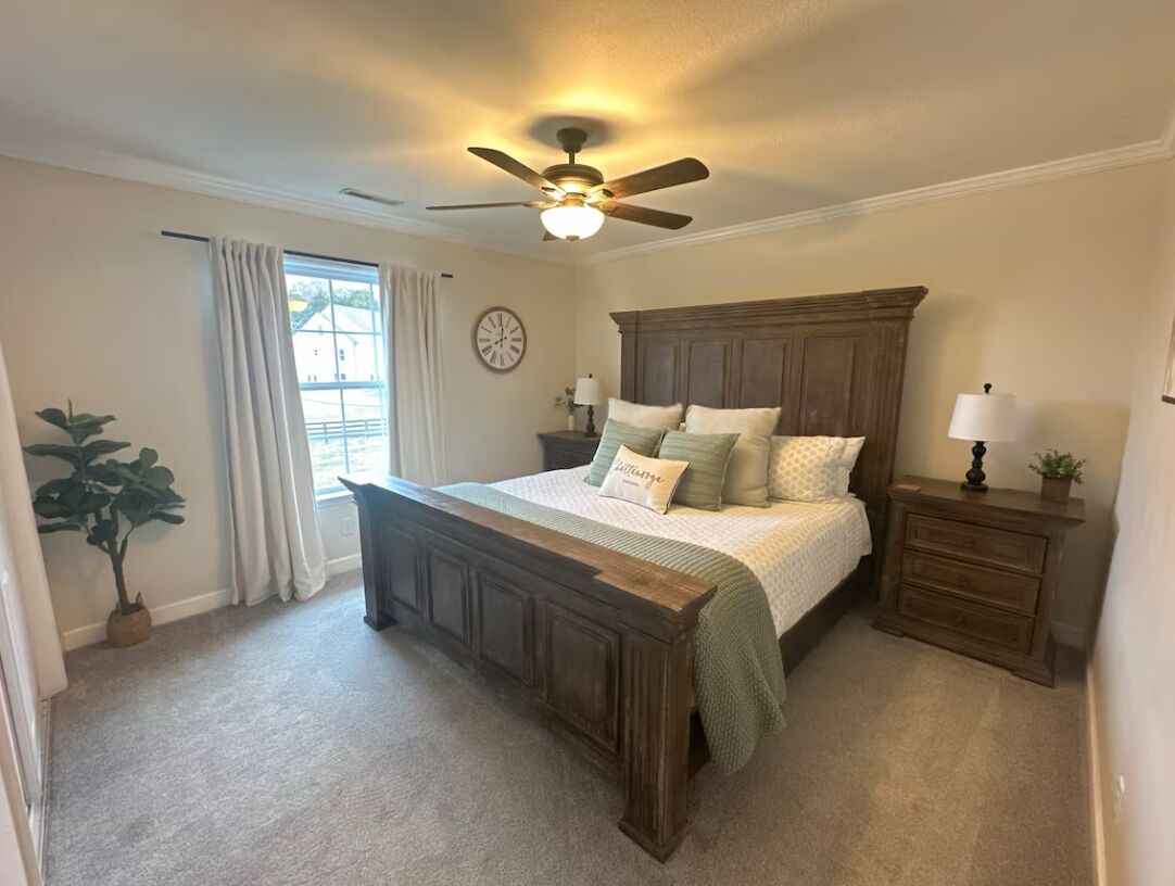 A bedroom with a wooden bed, a ceiling fan, a nightstand, a dresser, a potted plant, and a window with curtains.
