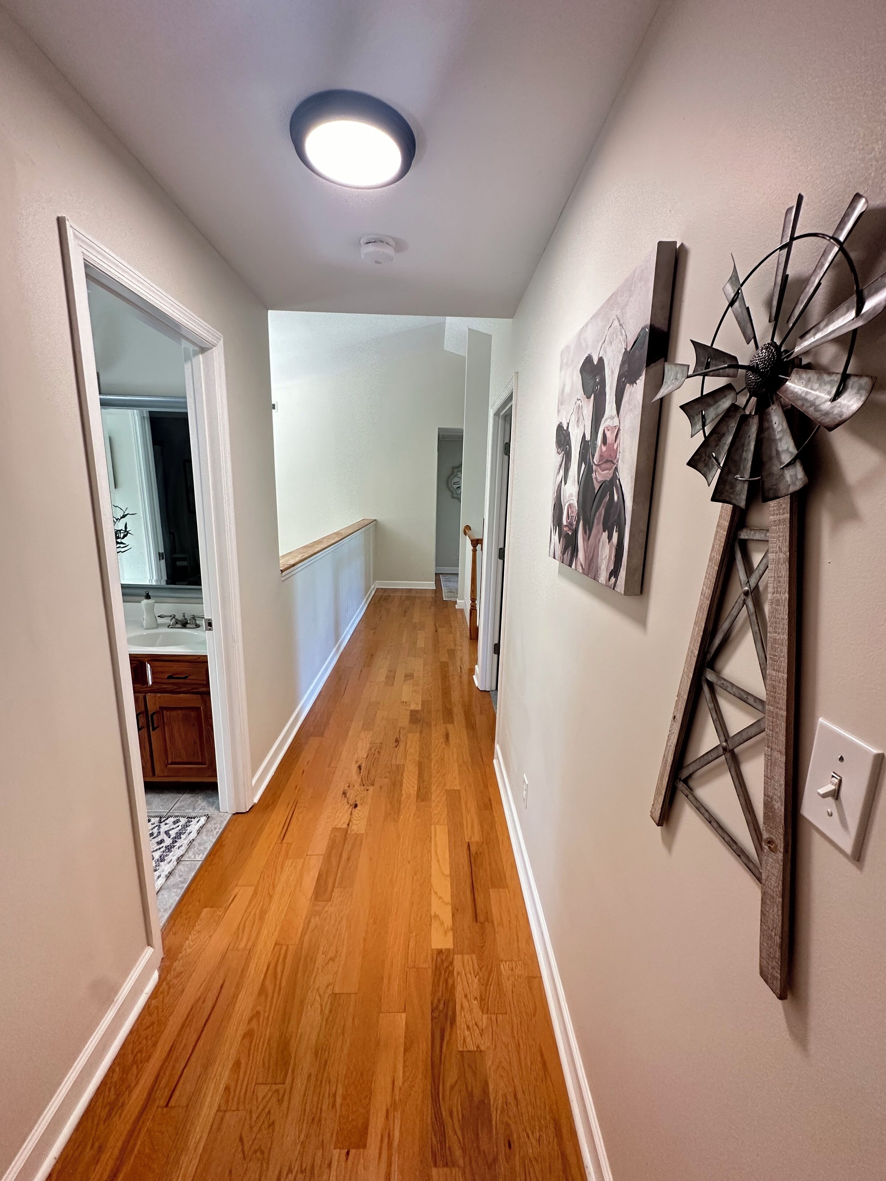 A hallway with wooden flooring, a ceiling light, a windmill decoration, and a painting of cows.