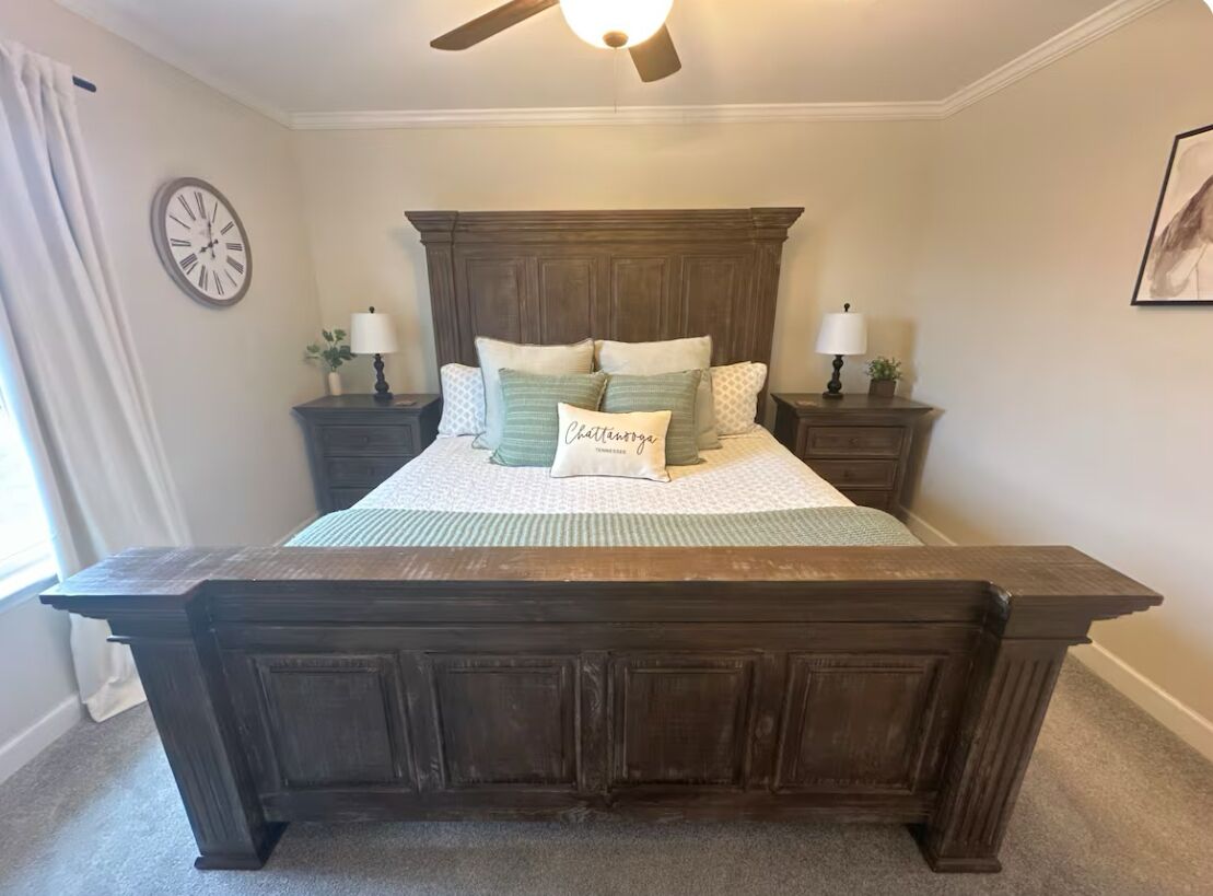 Quarto com uma cama grande, cabeceira de madeira, duas mesas de cabeceira com abajures, um relógio na parede e um quadro emoldurado.