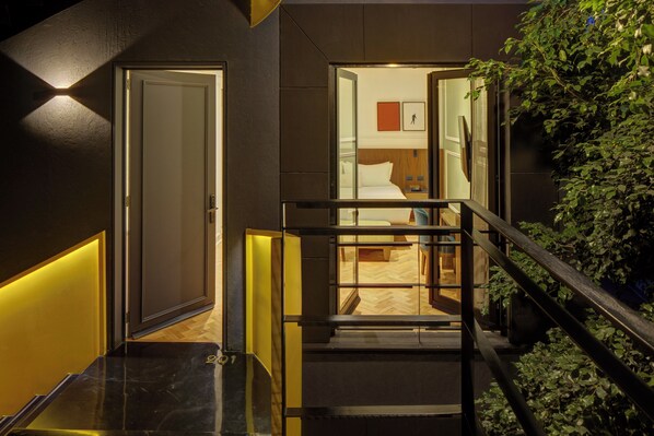 A modern hotel interior with a staircase, a visible bedroom with a bed and nightstand, and a wall-mounted picture.