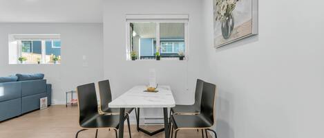 Een moderne eetruimte met een witte tafel en zwarte stoelen, een blauwe bank op de achtergrond, en een raam met planten.