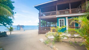 A two-story wooden building with a balcony, a beach view, and a small structure in the distance. - Modern Beachfront Casita with Dock and pool ON THE BEACH (Sandy Bay)