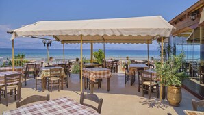 Un café en plein air avec des tables et des chaises disposées sous une pergola, offrant une vue sur la plage et la mer.