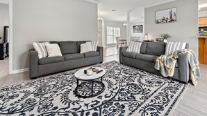 Un salon moderne avec deux canapés gris, un tapis à motifs et un coin repas à l’arrière-plan.