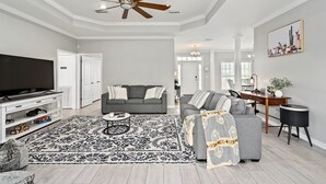 Un salon avec un ventilateur de plafond, une grande télévision, un canapé gris, un tapis à motifs et un bureau avec un ordinateur.
