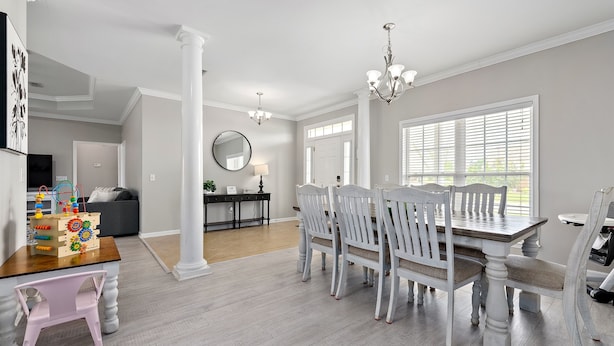 Un coin repas avec une table en bois et des chaises blanches, un lustre et une grande fenêtre avec des stores.